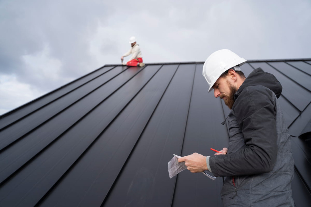 Roofing contractor in a hard hat taking notes while a second worker installs a metal roof in the background