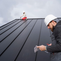 Roofing contractor in a hard hat taking notes while a second worker installs a metal roof in the background