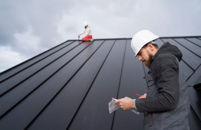 Roofing contractor in a hard hat taking notes while a second worker installs a metal roof in the background